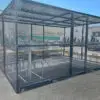 A secured outdoor bike storage cage, featuring customizable bike sheds and multiple bike racks inside, is located in an industrial area with warehouses in the background. The 40 Space Two Tier Security Cycle Enclosure has metal mesh walls and a roof, providing protection and security for bicycles.