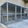 An outdoor metal cage structure designed for secure bicycle storage, the 40 Space Two Tier Security Cycle Enclosure features multiple individual racks and sturdy wire mesh walls. Situated next to a brick building on a paved surface, it offers customisable locking options for enhanced security.