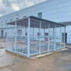 A 40 Space Two Tier Security Cycle Enclosure with a corrugated roof is situated beside a large, white industrial building. The shed is enclosed with wire mesh and has several cycle spaces inside. The ground around the enclosure is wet, suggesting recent rain and highlighting multiple locking options available.
