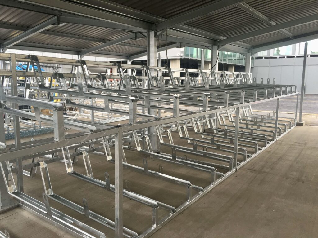 A covered bike storage facility with multiple rows of vertical bike racks. The metallic structure is empty, and the surrounding area is partially visible under the canopy roof. The facility is located in an urban environment.