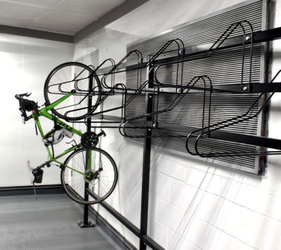 A green road bike is hanging vertically by its front wheel on an industrial-style bike rack attached to a white brick wall. The bike rack is made of black metal and includes several curved hooks for multiple bicycles. The flooring is gray and the setting appears to be a garage or storage area.