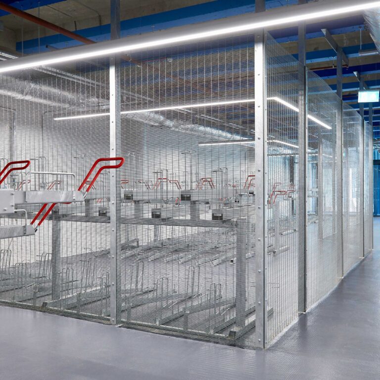 A large, well-lit room containing several rows of metal two tier bike racks enclosed by the anti-climb mesh fencing. The ceiling features exposed pipes and beams.