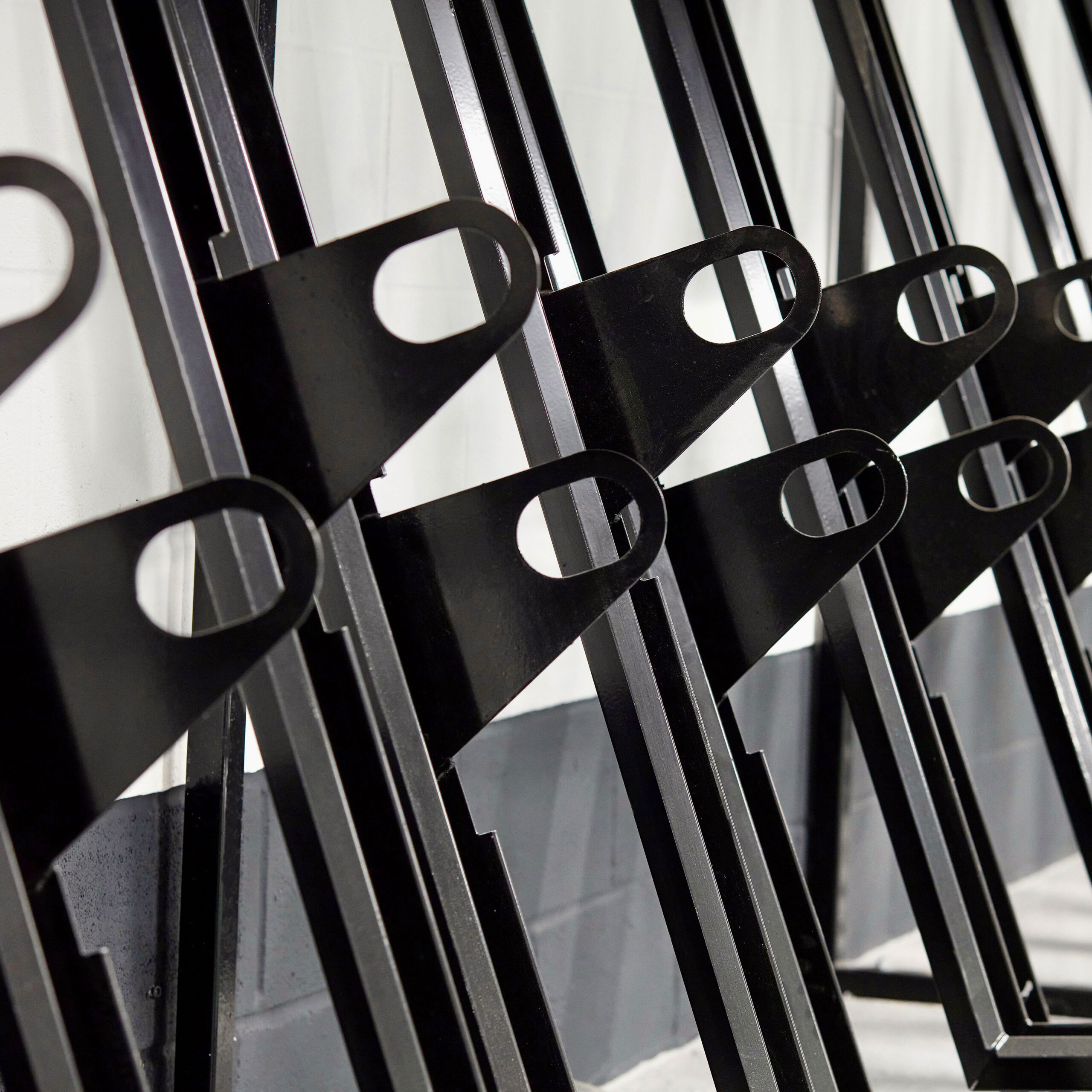 Close-up of several black metal semi vertical bike racks positioned in a row. The brackets have a sleek, modern design with a circular cutout at one end. They are arranged diagonally against a white and gray background.
