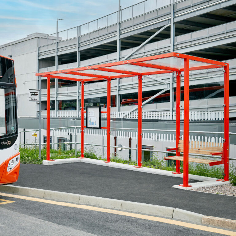 A modern bus shelter with a bright red frame and a transparent roof, located near a multi-story parking garage. A bench is inside the shelter, and a bus is parked nearby. The sky is partly cloudy with visible blue patches.